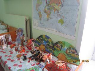 На фоне карты мира на столе представлены экспонаты: веера, куклы, шляпы, сувениры.